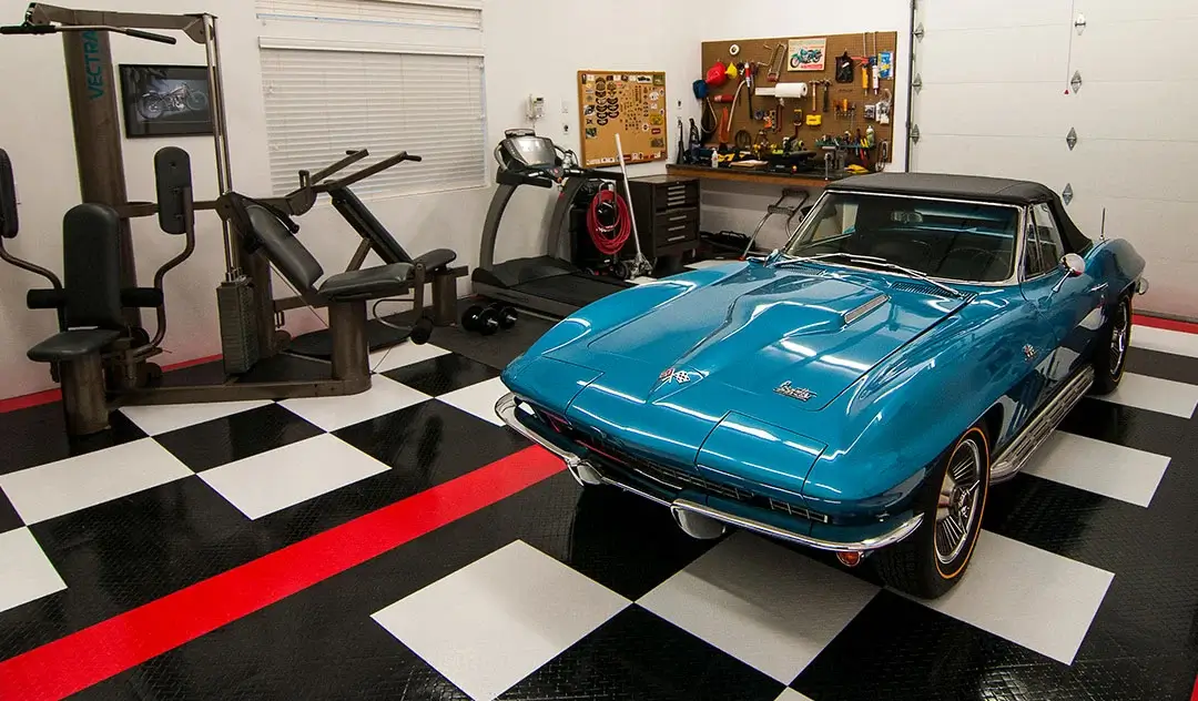 Vintage Corvette parke in a garage with gym equipment. The garage has a checkerboard pattern with a red stripe on its garage flooring tiled floor.